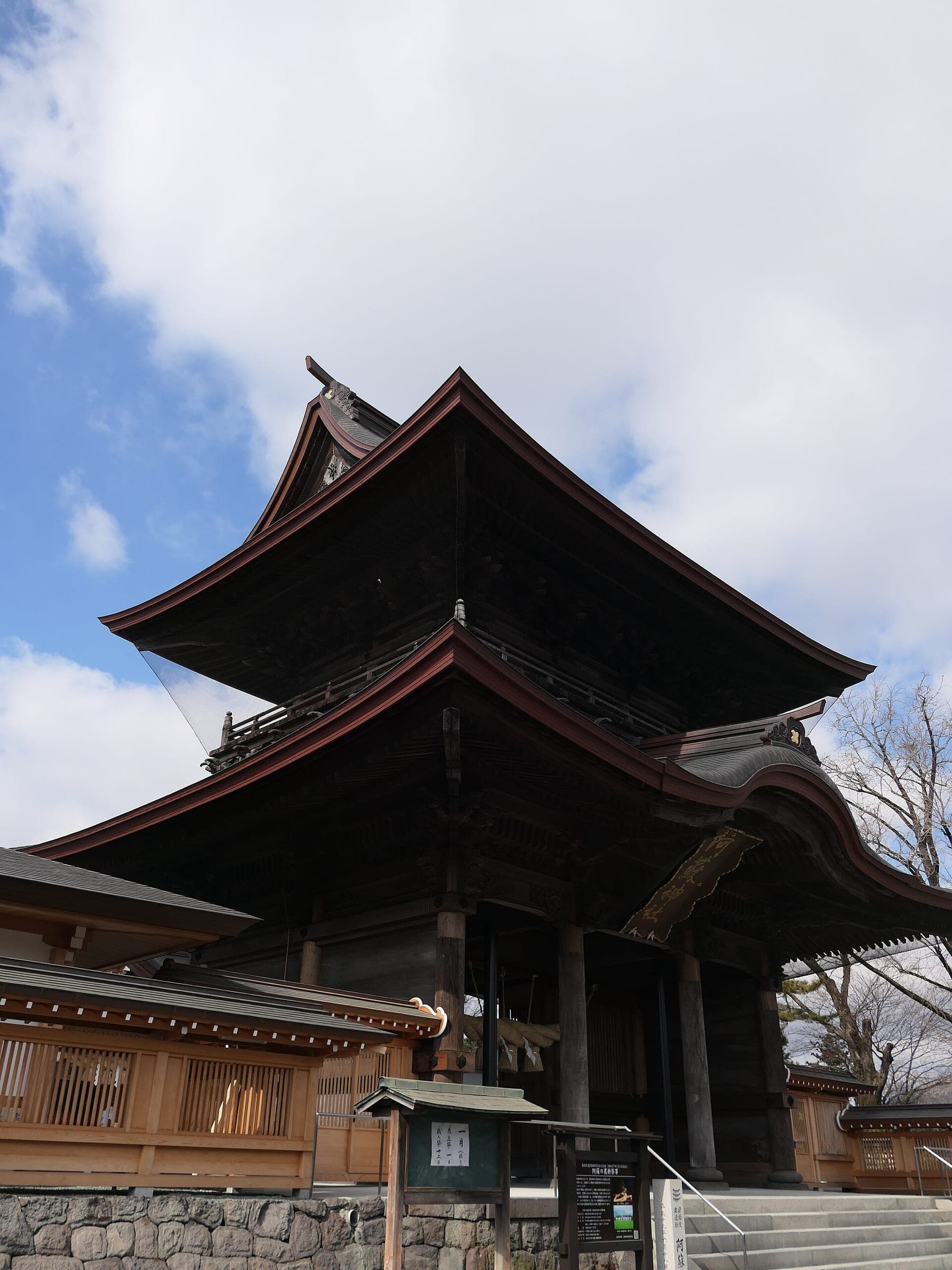 The two-storey Romon gate of Aso Shrine rises in dark timber under a blue sky in Ichinomiya.