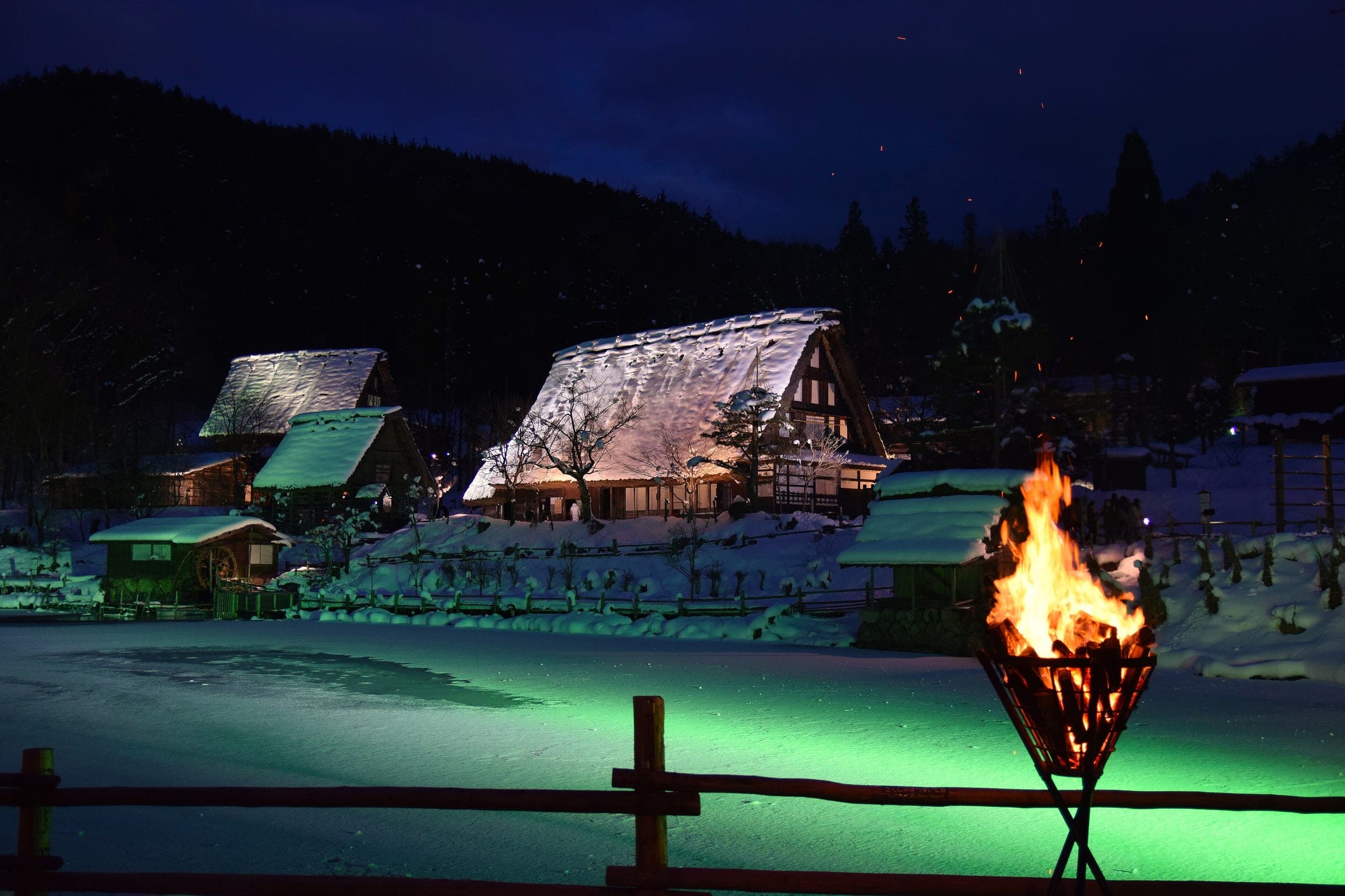 Lights and a fire basket glow below snow-covered thatched farmhouses at Hida Folk Village on a winter night