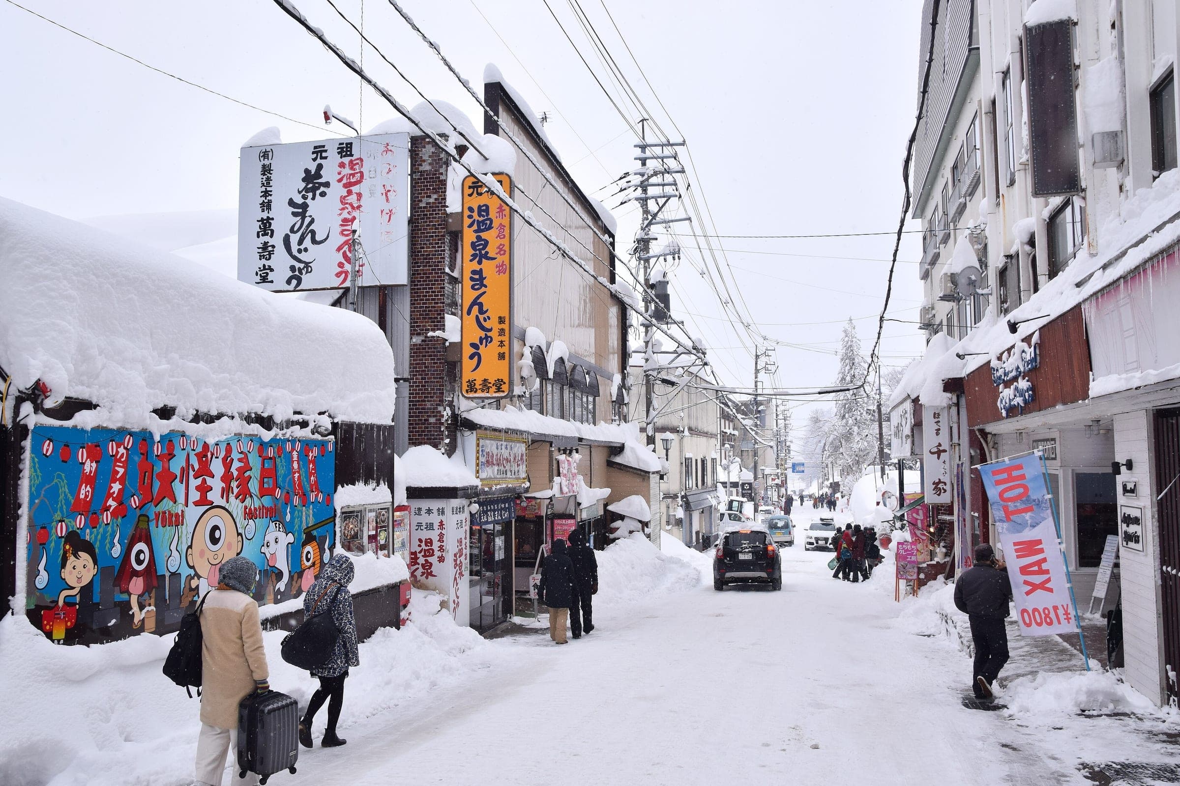Visitors with luggage walk Akakura Onsen's snowy main street past inns and souvenir shops in Myoko