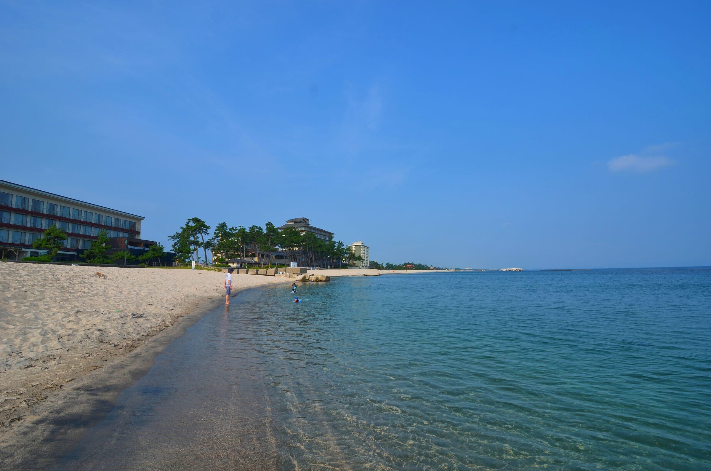 Clear water laps the long sandy beach below the hotels of Kaike Onsen in August