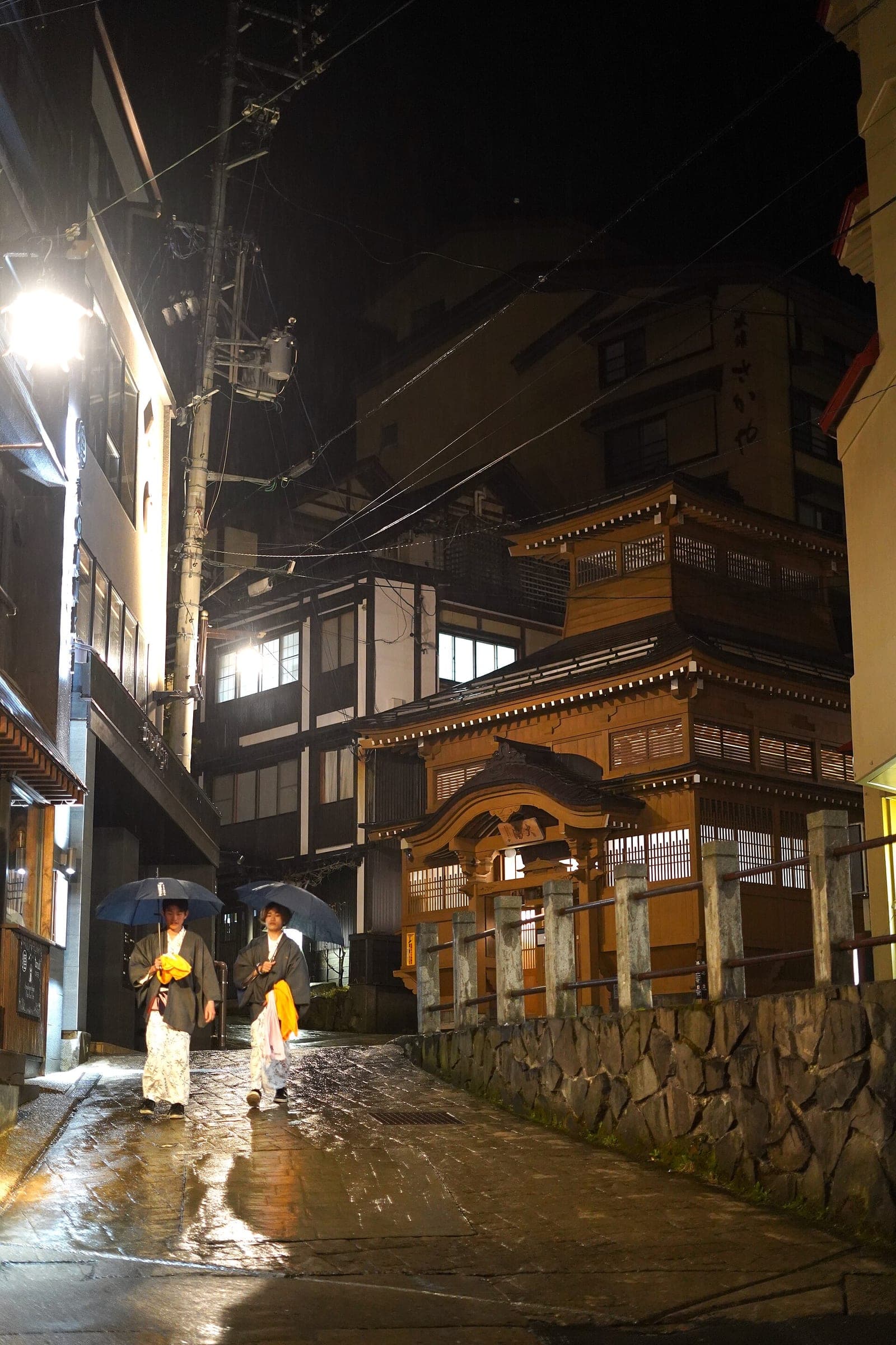 Two bathers in yukata walk past the lit Oyu bathhouse in Nozawa Onsen on a rainy night.