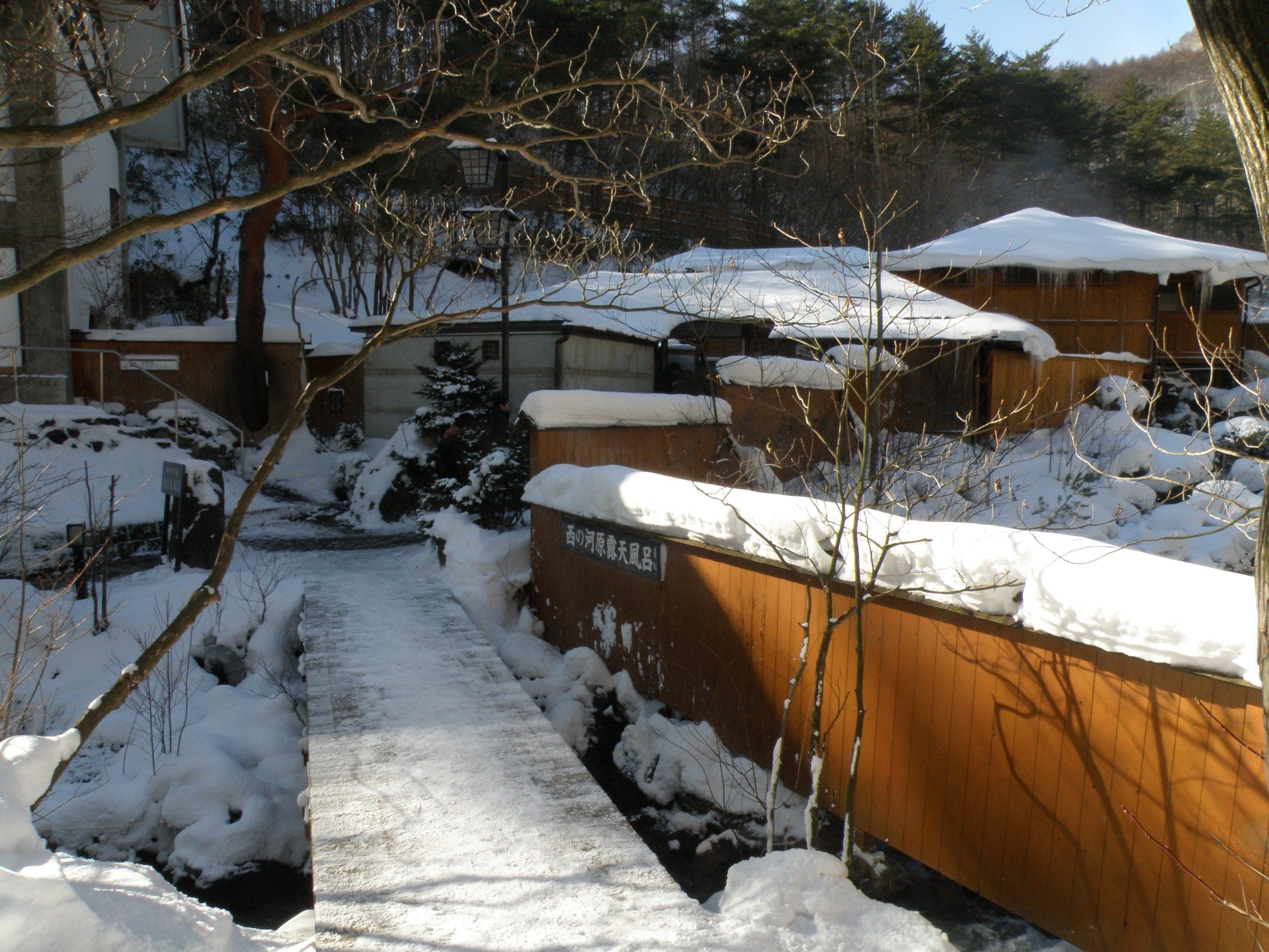 A snowy boardwalk leads past wooden fences to the Sai-no-kawara Rotenburo in Kusatsu