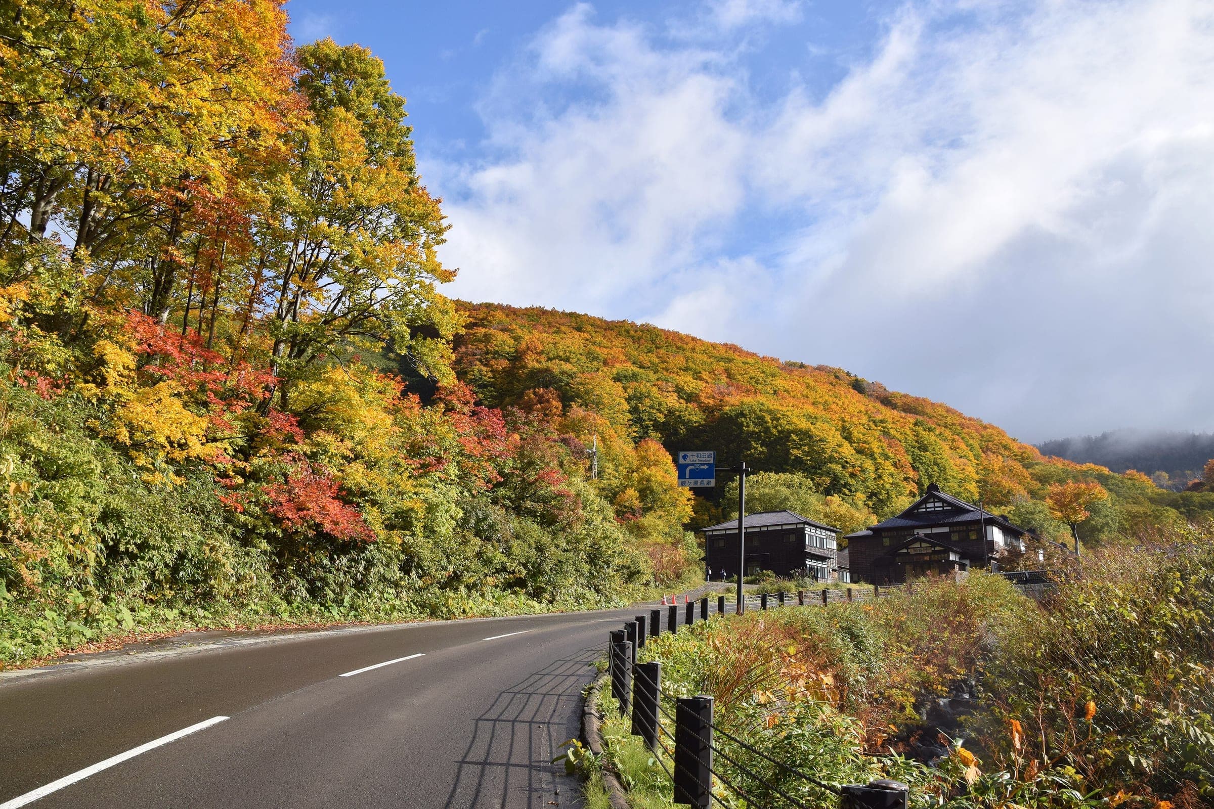 The road climbs past the dark timber buildings of Sukayu Onsen through bright October foliage.