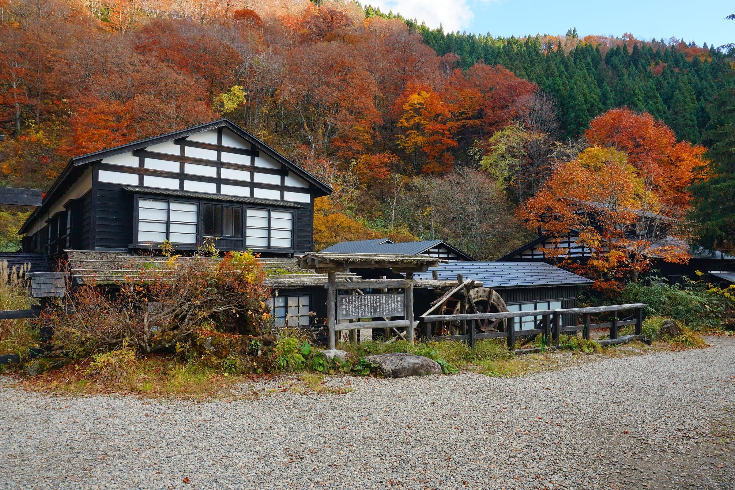 Black timber lodgings and a water wheel sit below red autumn hills at Tsurunoyu Onsen.