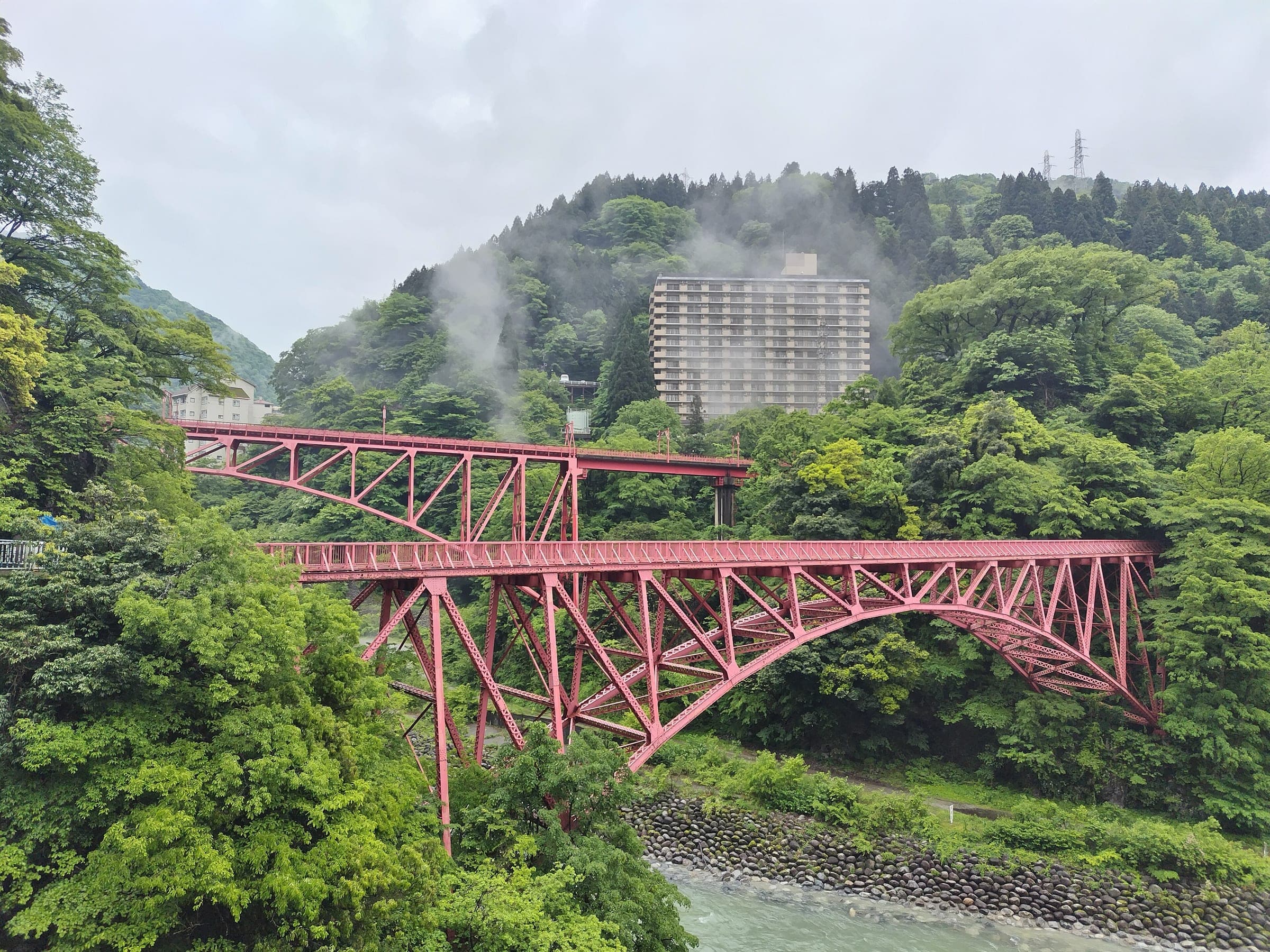 Steam drifts from the hillside behind the red Yamabiko bridges and a hotel at Unazuki Onsen