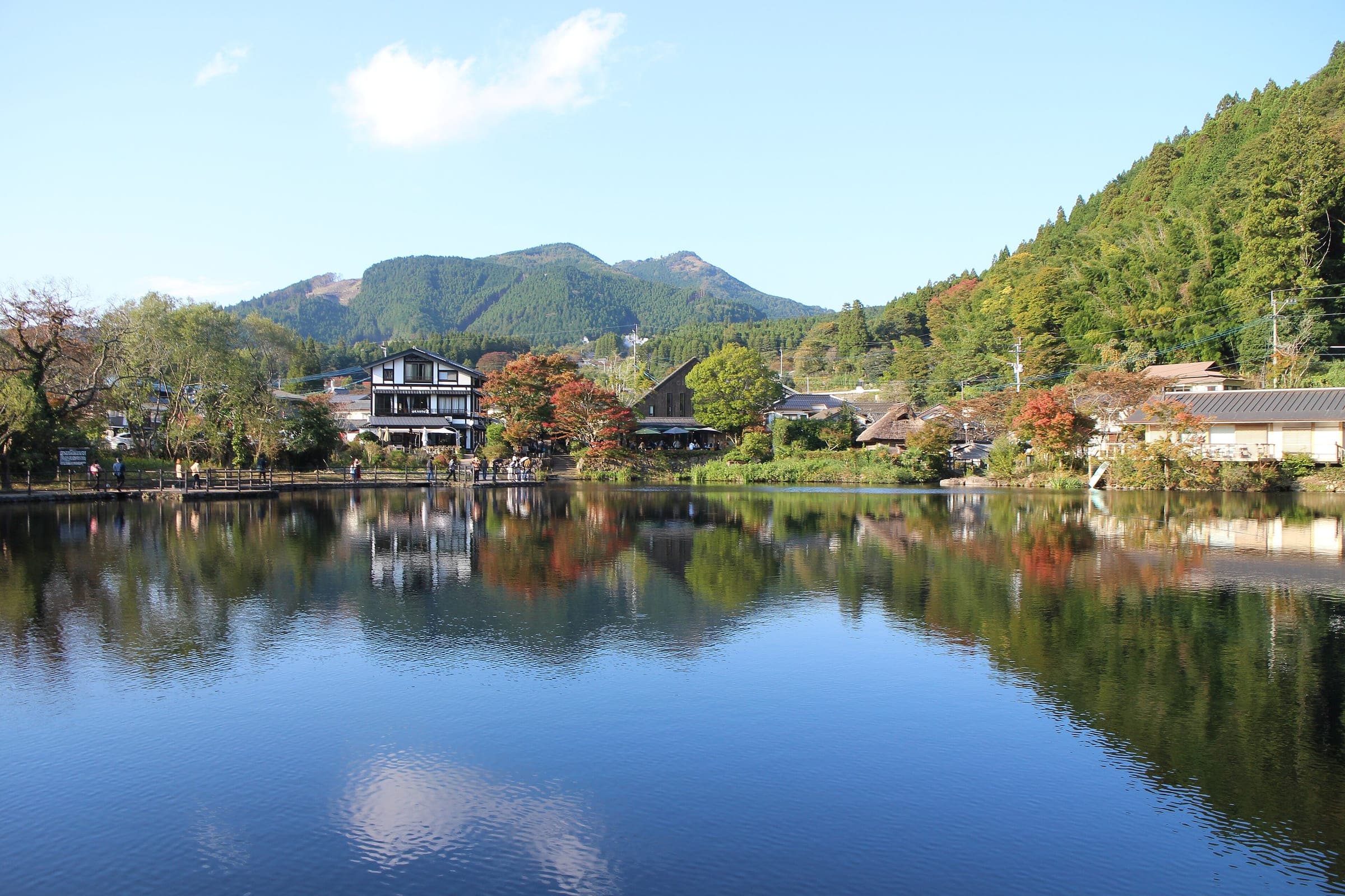 Autumn trees and Yufuin's houses reflect in the calm surface of Lake Kinrin under a blue sky.