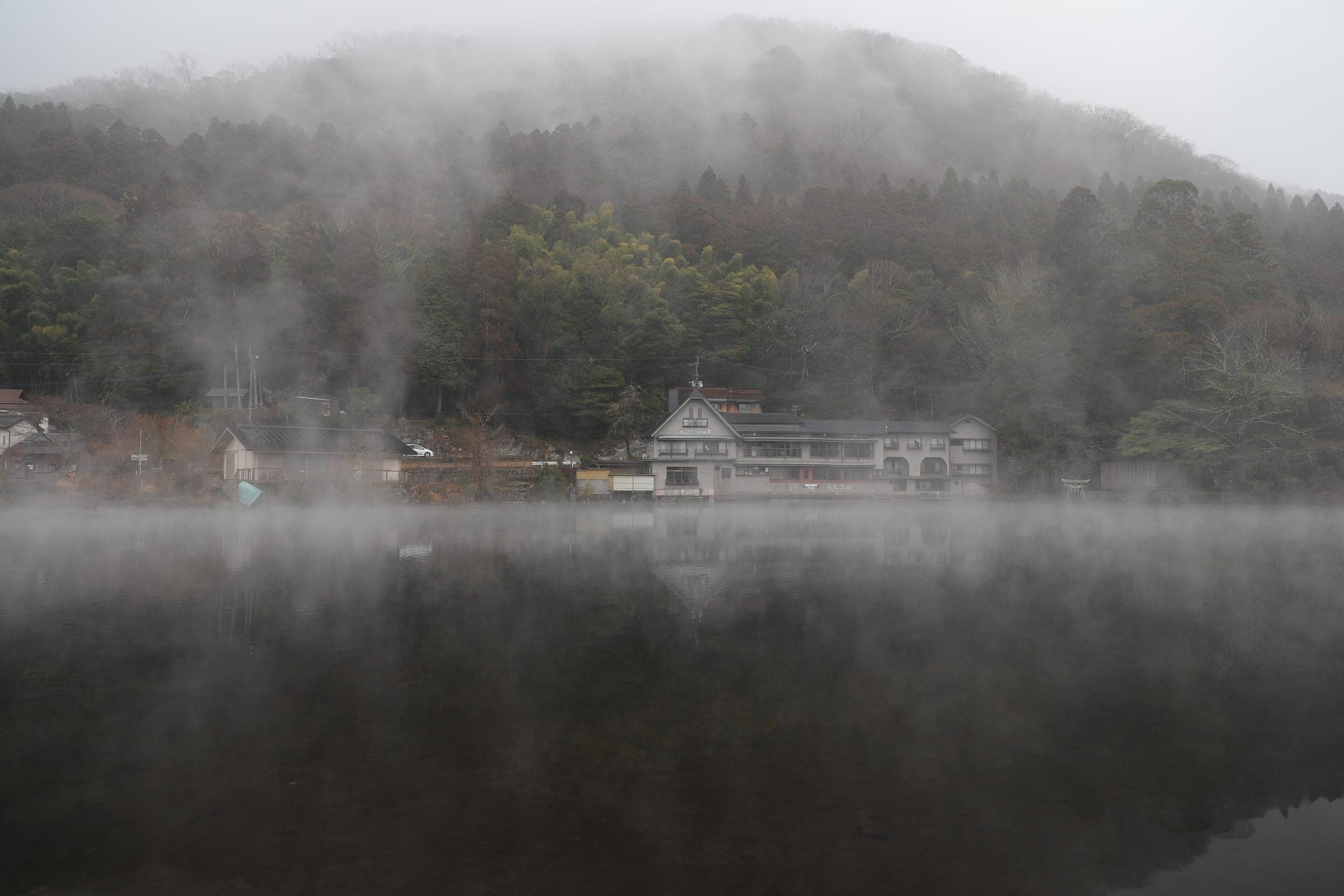Fog hangs over the still water of Lake Kinrin and the wooded hill behind it in Yufuin.