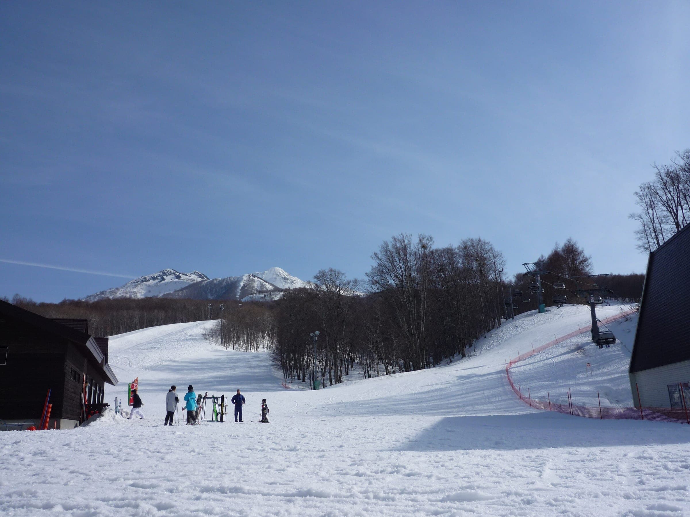 Skiers gather at the base of Aomori Spring with the white peaks of Mount Iwaki behind the trees.