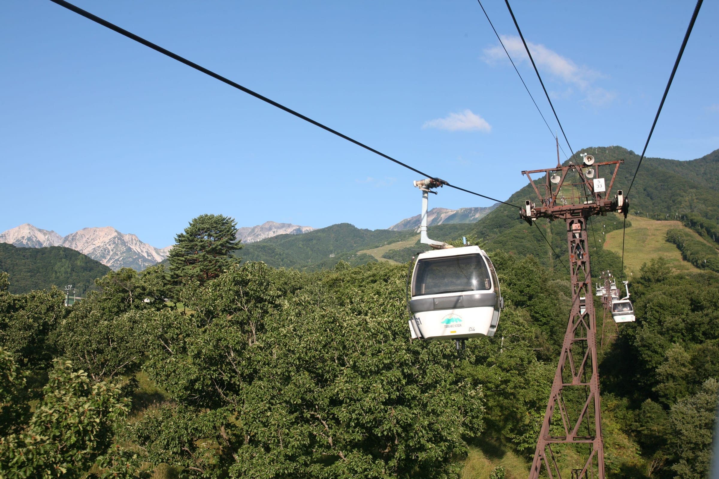 A Tsugaike gondola cabin rises over green forest toward the Hakuba peaks in late summer