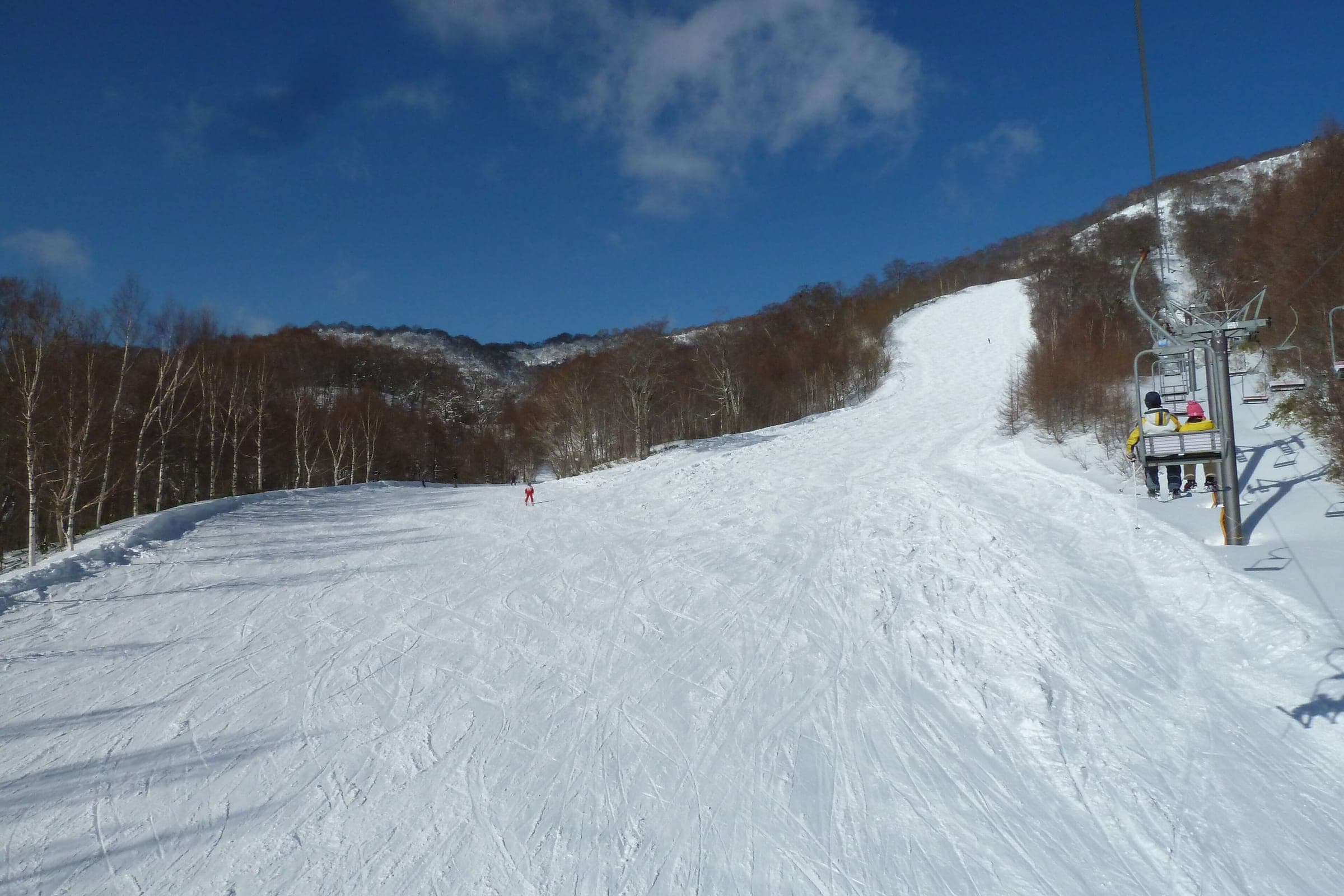 Skiers ride the Nishiyama lift beside the Mizunara run at Oze Iwakura under a blue sky