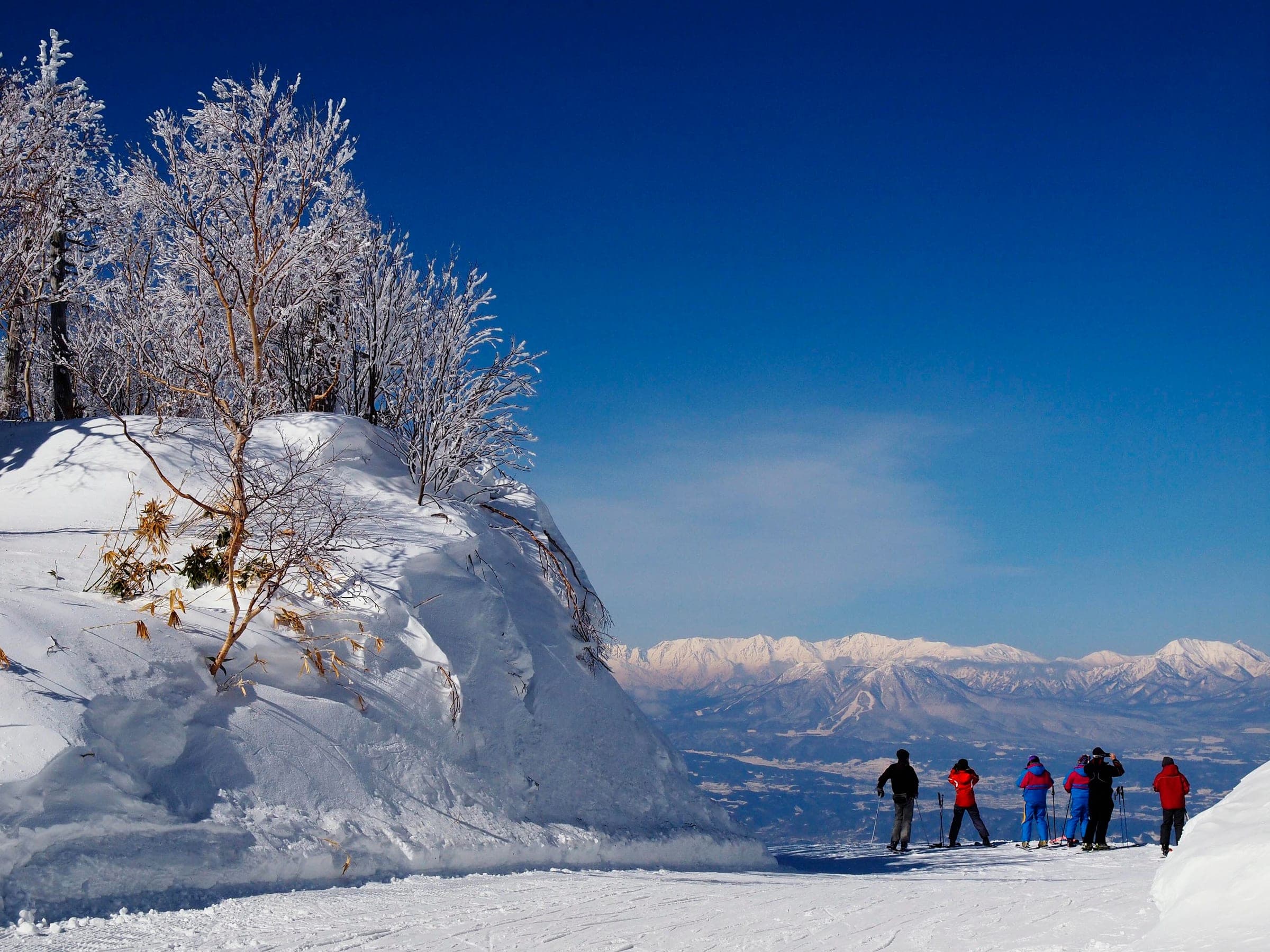 Skiers pause on a groomed run at Shiga Kogen Takamagahara as the Northern Alps rise white across the valley