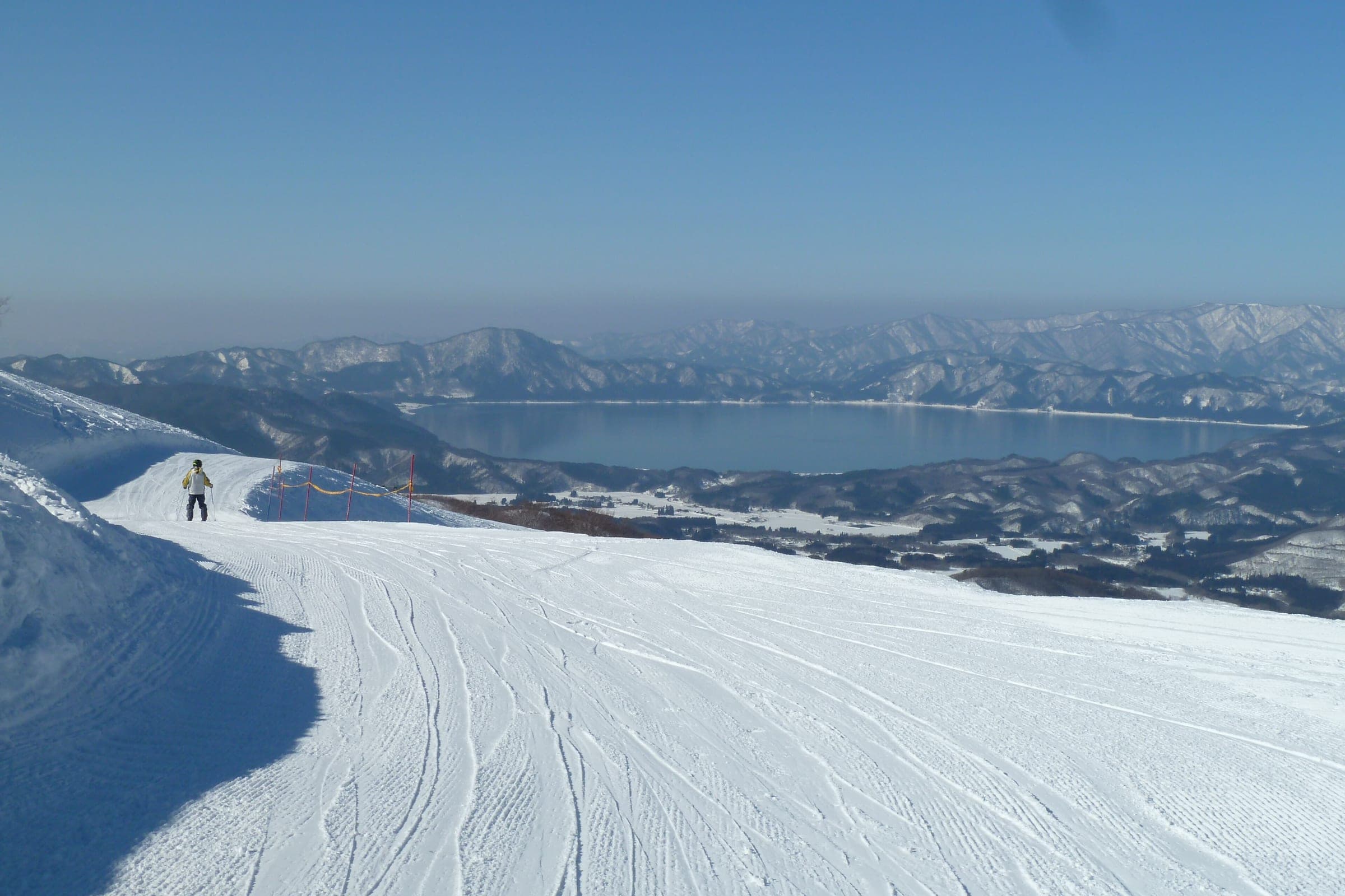A skier pauses on fresh corduroy at Tazawako ski area with Lake Tazawa spread out below.