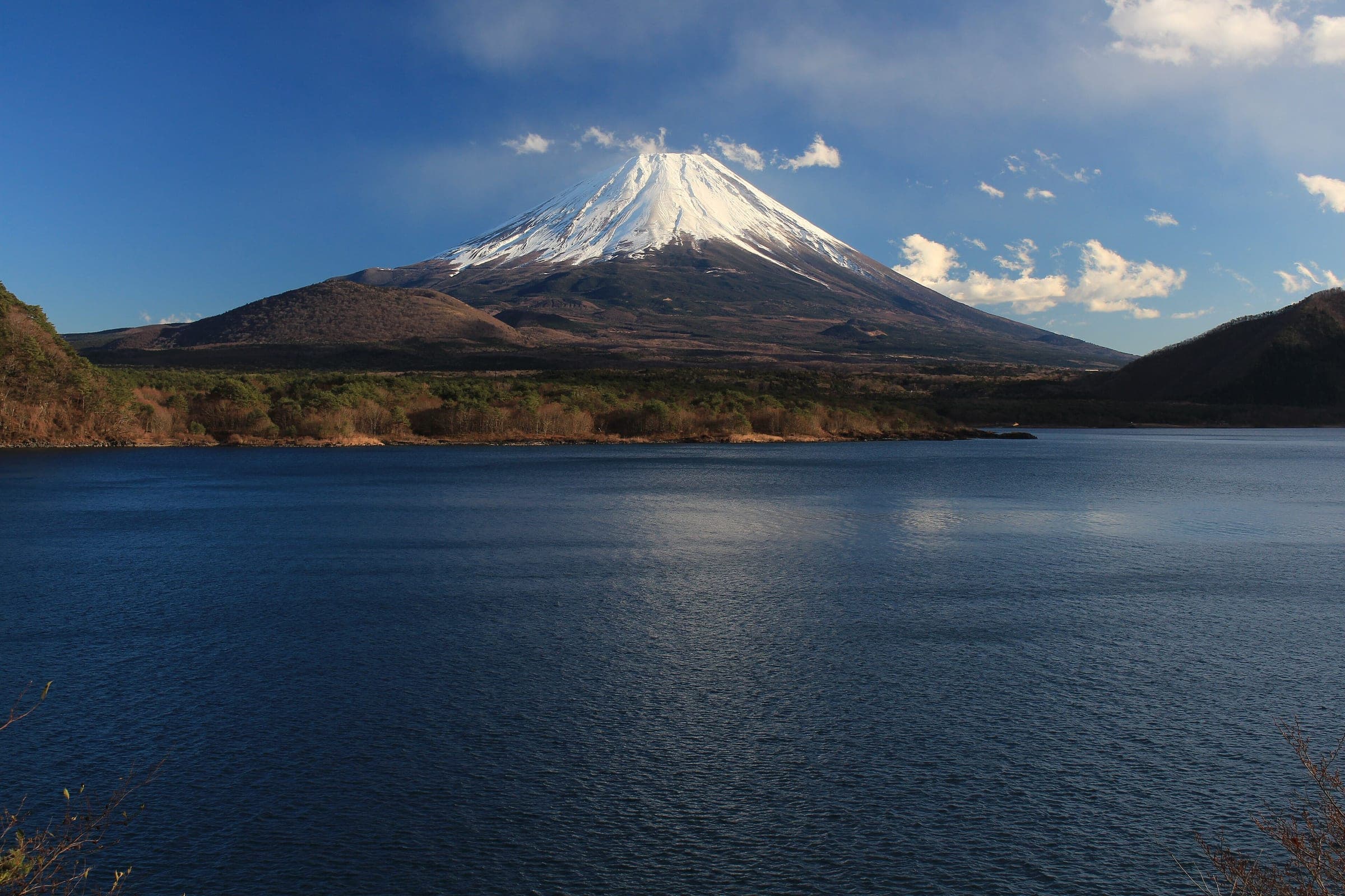 Mt. Fuji rises with a snowy summit above the deep blue water of Lake Motosu.
