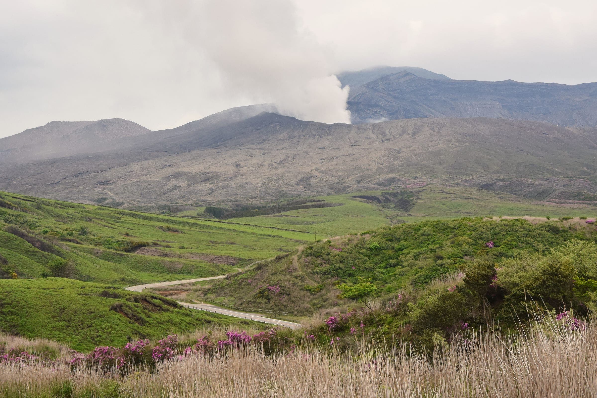 Steam rises from the Nakadake crater above green slopes and a winding mountain road on Mount Aso.