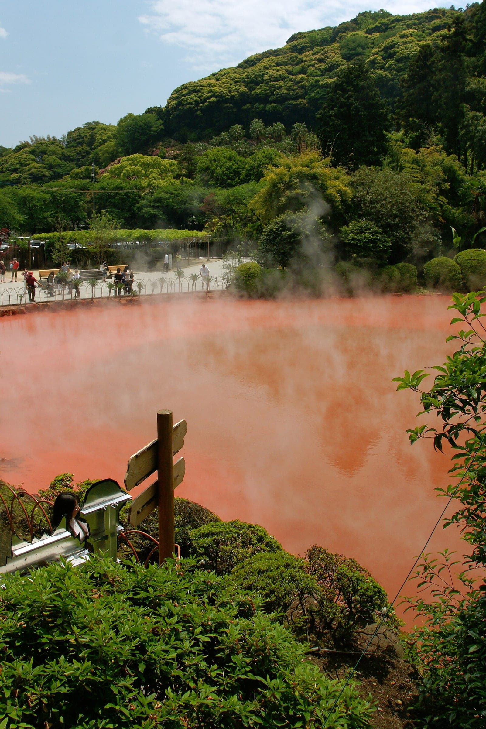 Mist drifts over the red clay water of Chinoike Jigoku below green hills in Beppu.