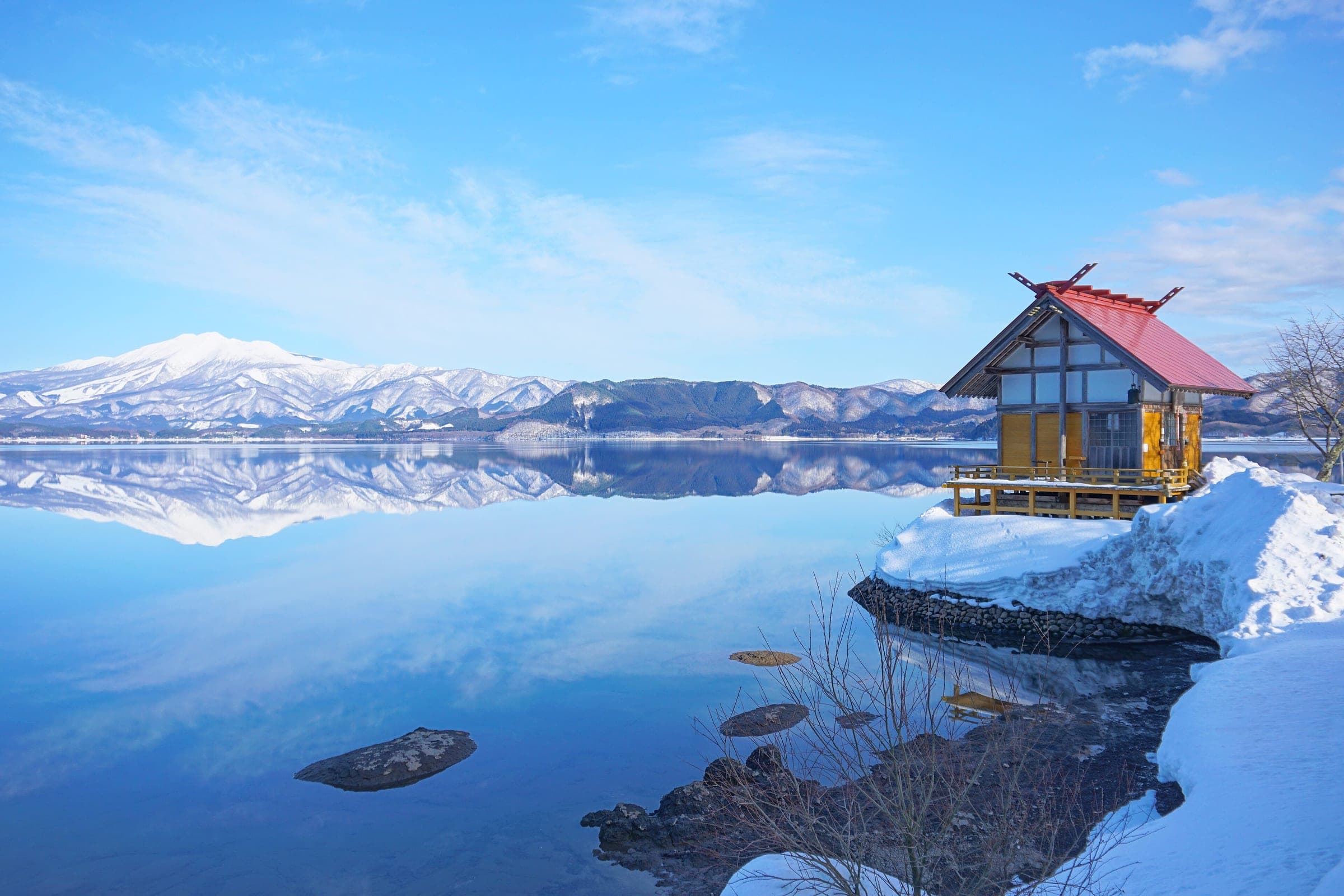 The red-roofed Kansa-gu stands on a snowy point of Lake Tazawa as white peaks mirror in still water.