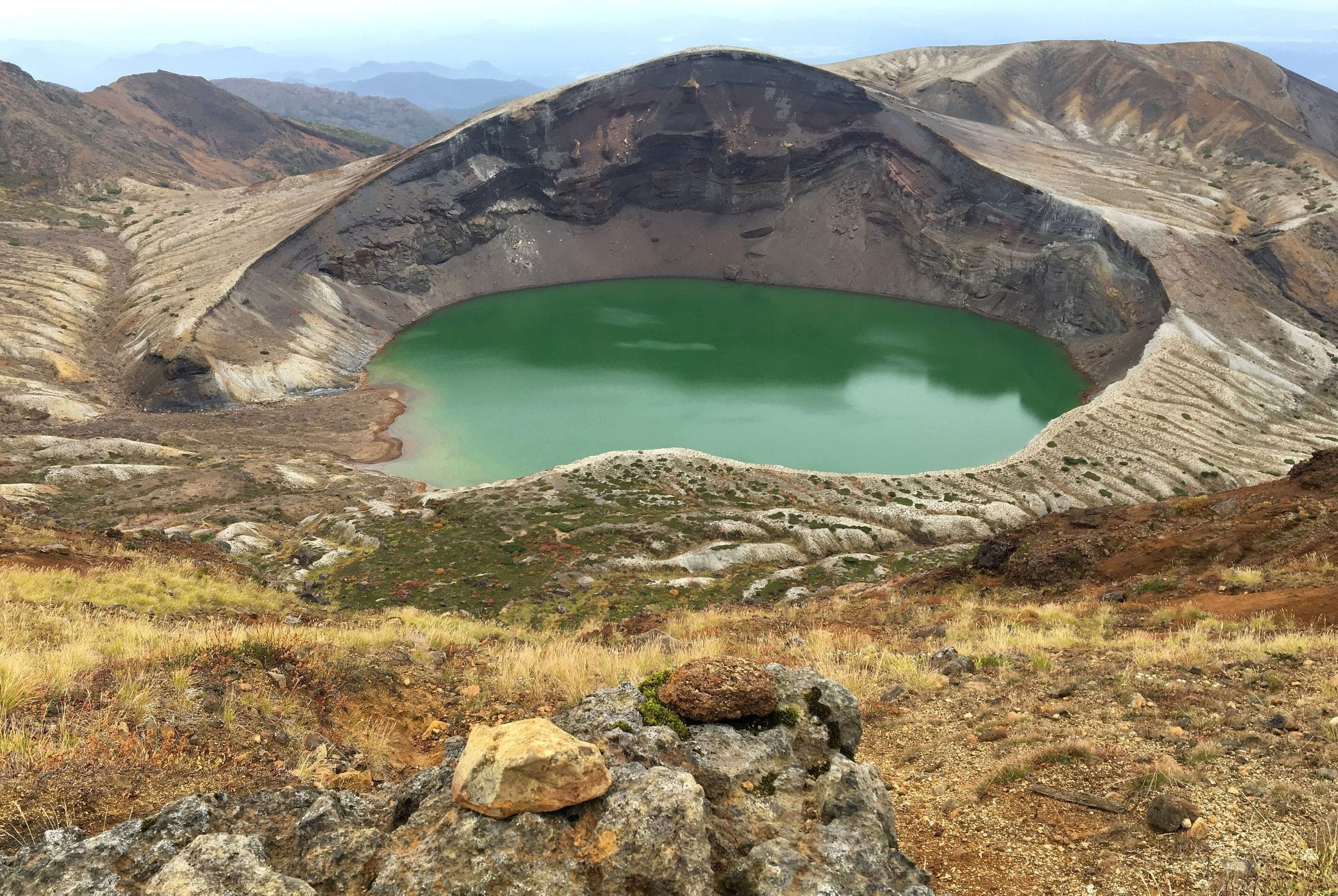 The green crater lake of Okama sits inside banded volcanic walls near the summit of Mount Zao.