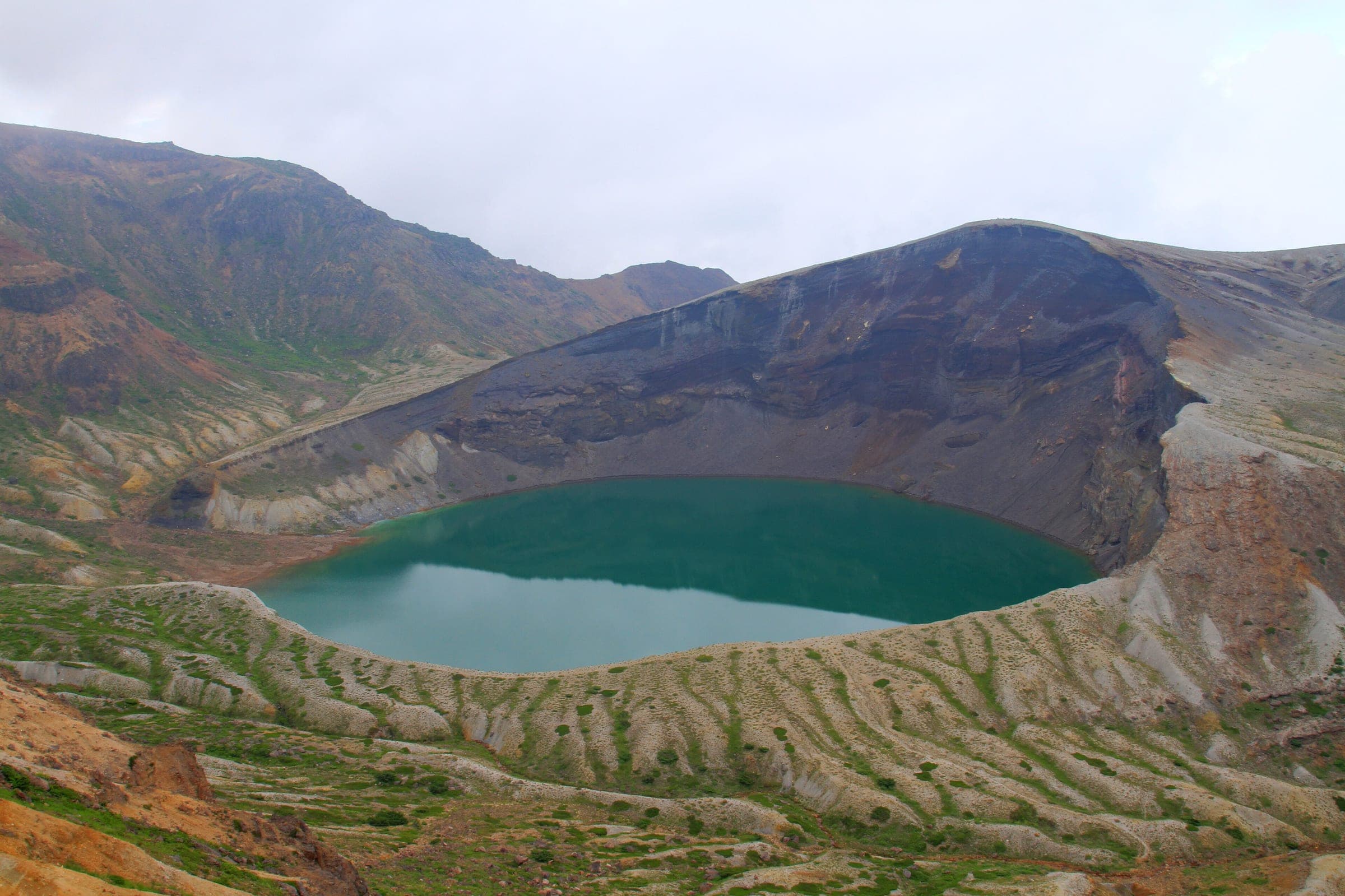 Okama crater lake lies below its dark inner wall on Mount Zao under a soft grey sky.