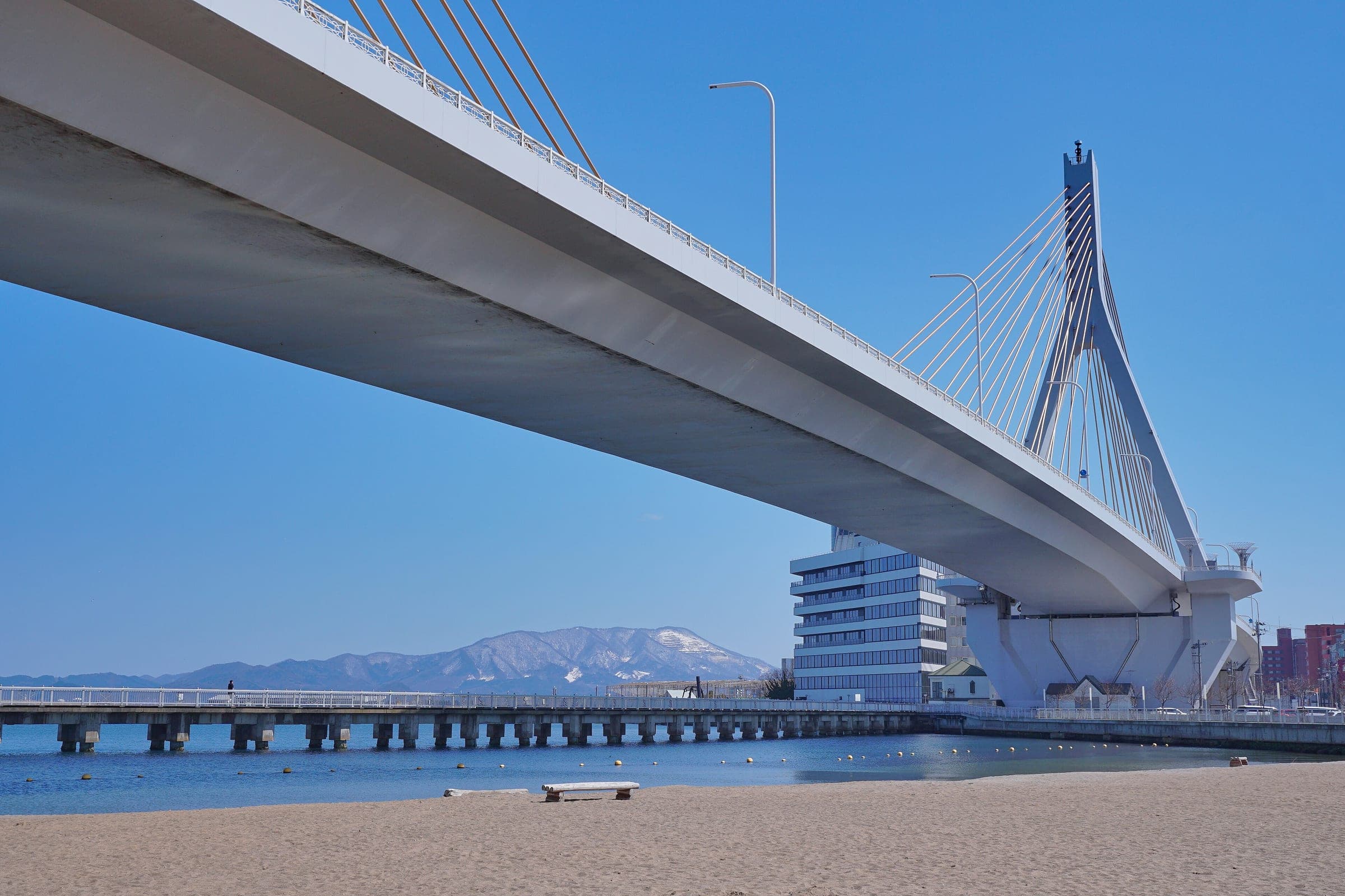 Aomori Bay Bridge arcs over a small city beach with snowy mountains across the bay in March.