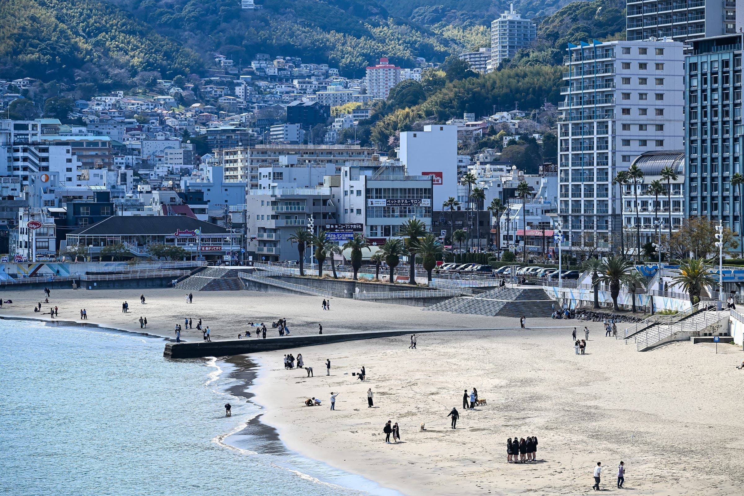 Walkers cross the sand of Atami Sun Beach below the palm-lined promenade and hotel towers.