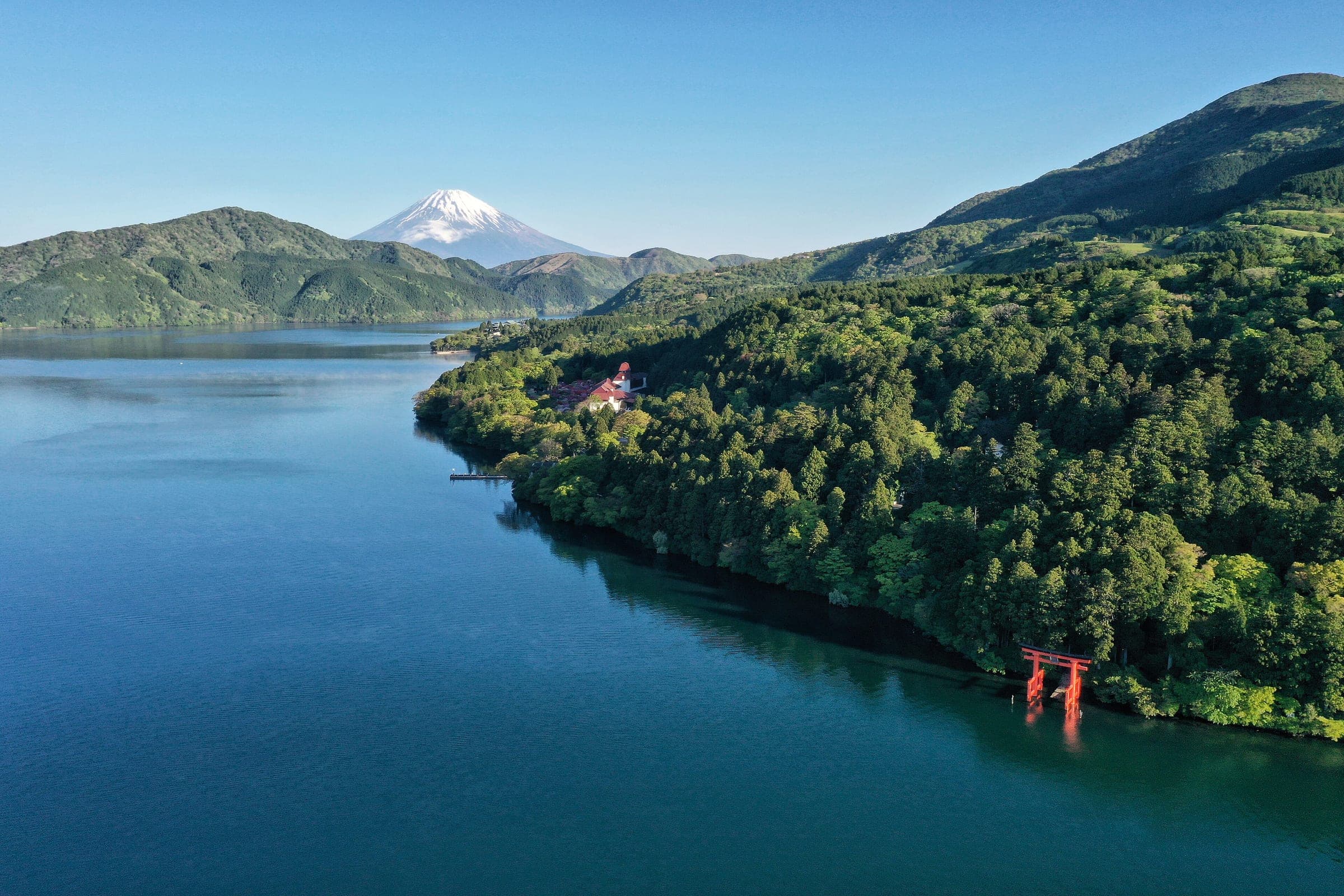 The red torii of Hakone Shrine stands in Lake Ashi below forested slopes as Mt. Fuji rises on the horizon.