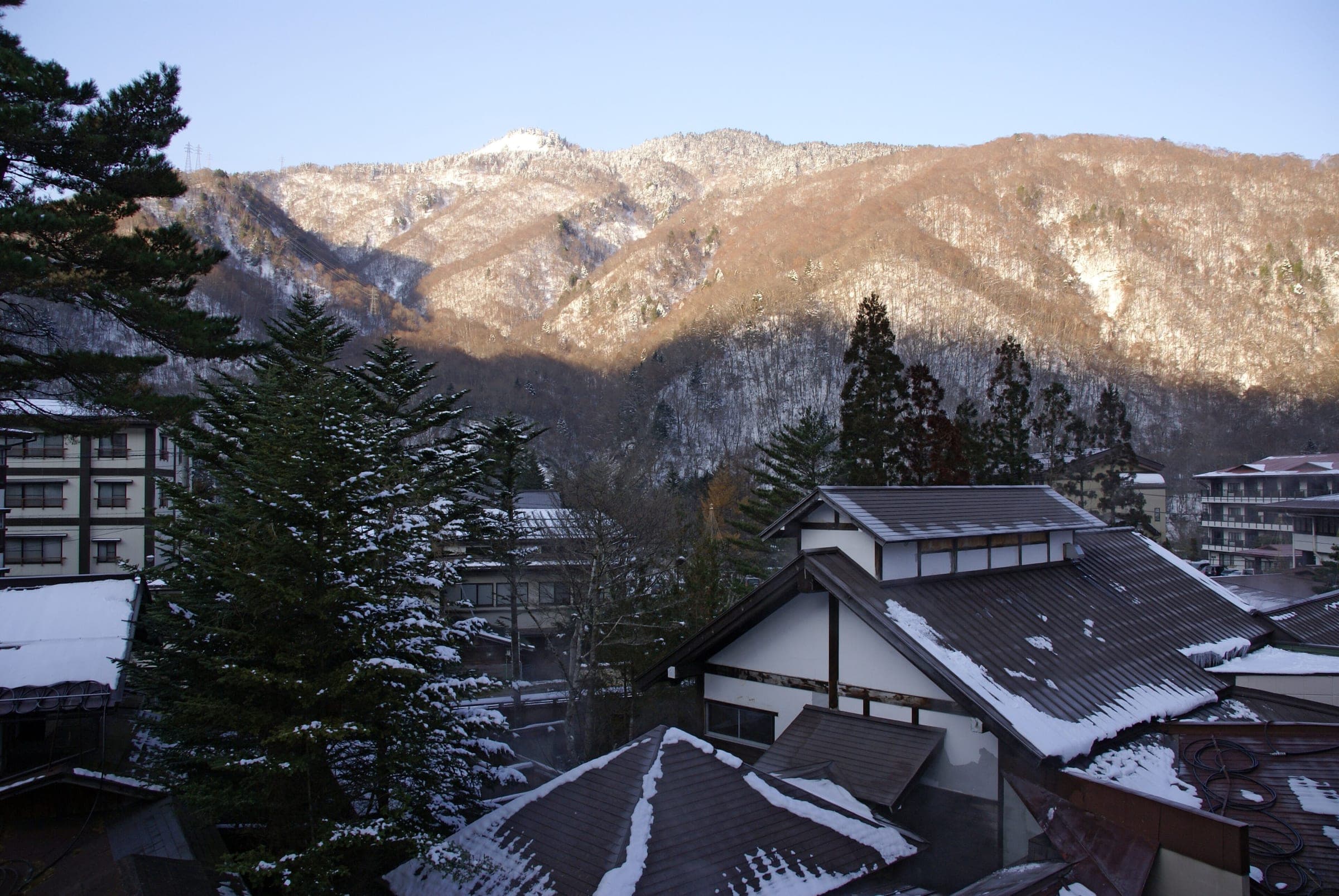 Fresh snow settles on the inn rooftops of Hirayu Onsen below a sunlit mountainside in late November