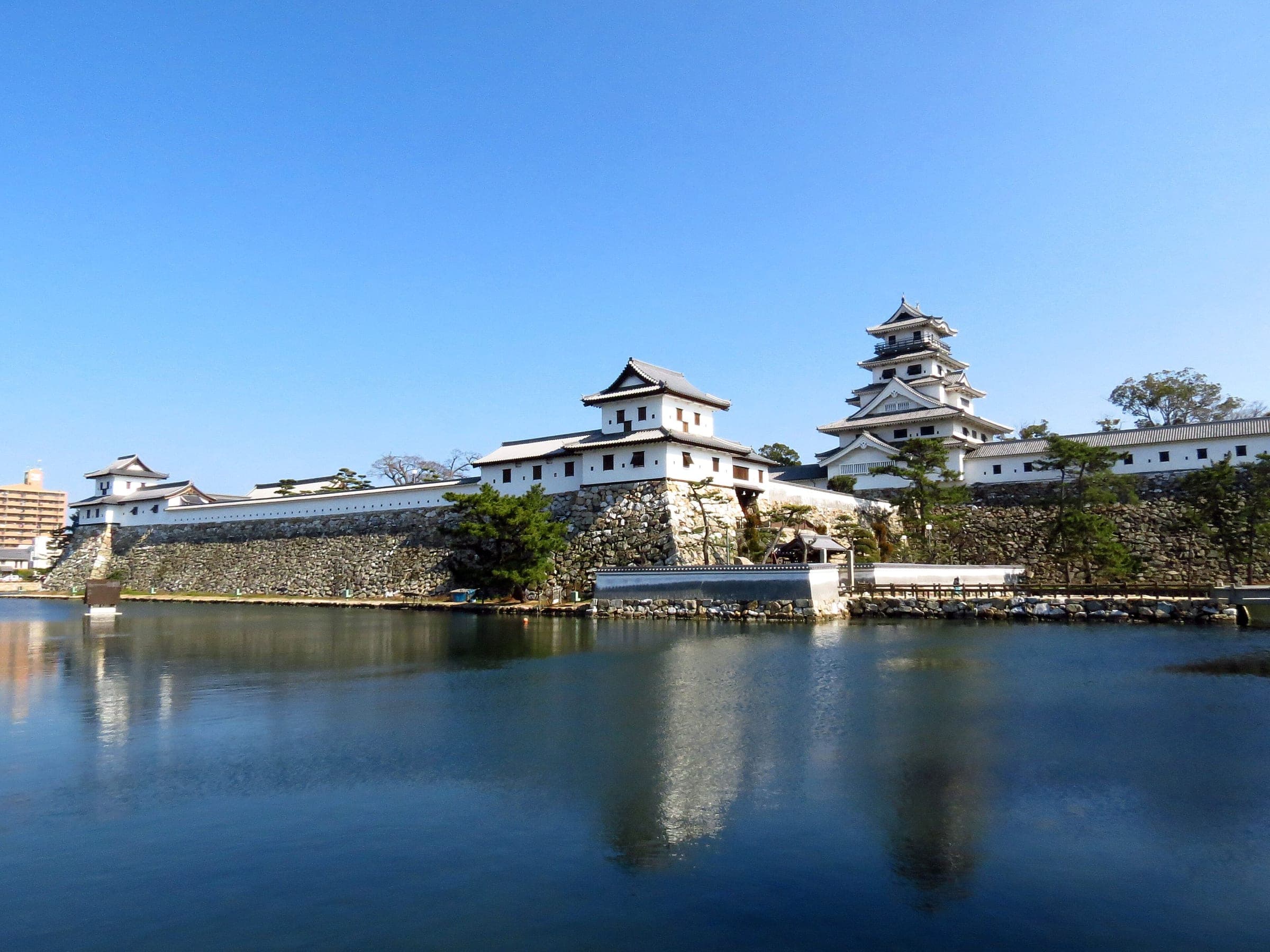 Imabari Castle's white keep and turrets reflect in its broad seawater moat under a clear sky.
