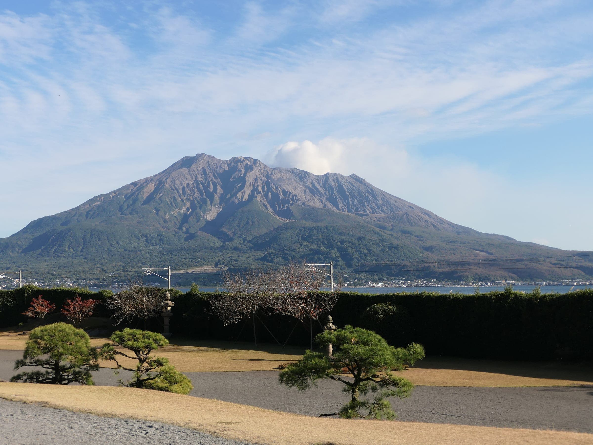 A small plume rises from Sakurajima across Kagoshima Bay beyond the lawns and pines of Sengan-en.