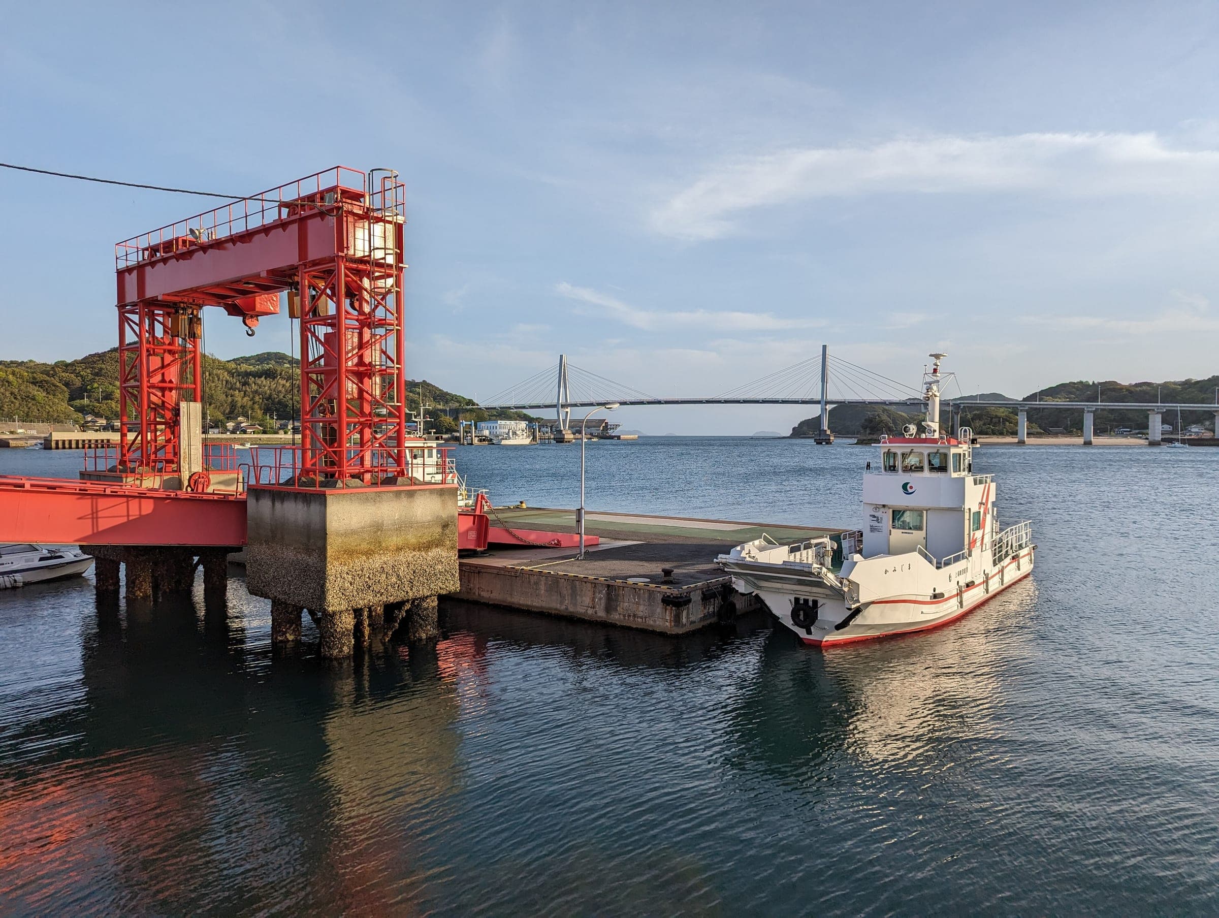 A small ferry waits at the red pier on Yugeshima, with the Yuge Bridge across the water in Kamijima.