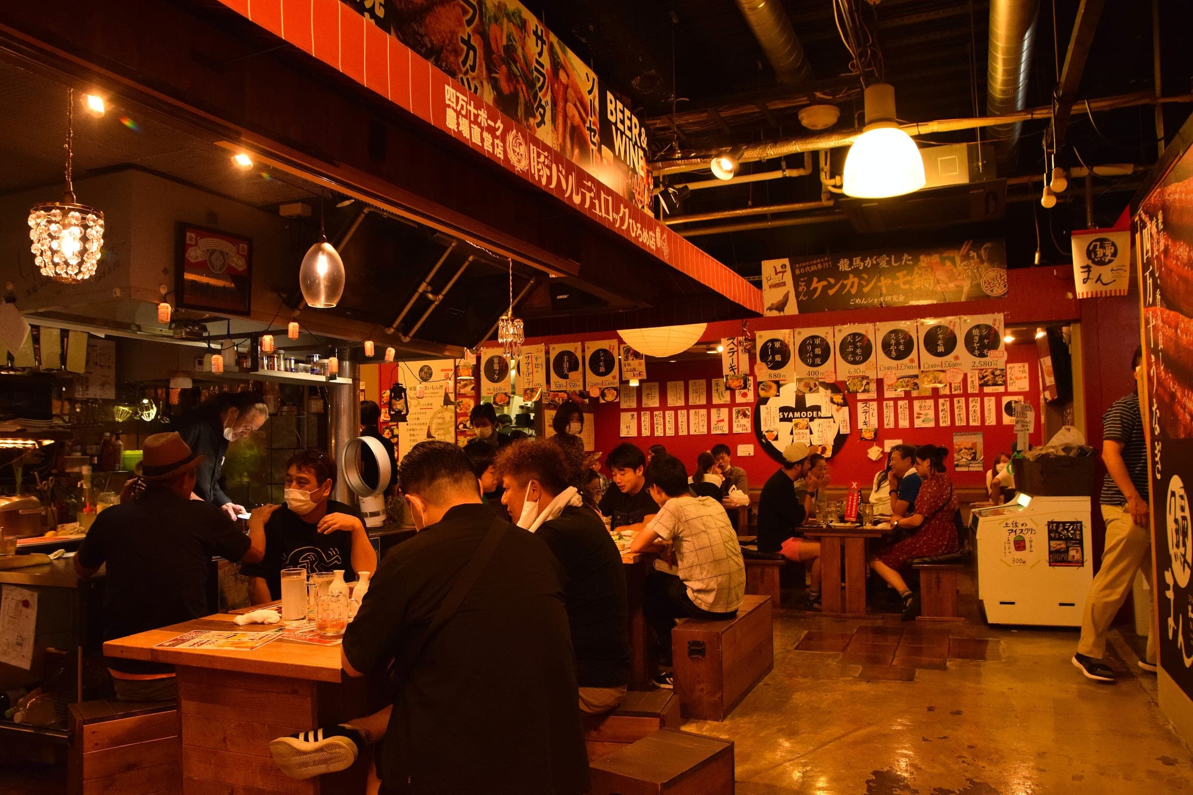 Diners crowd the long wooden tables of Hirome Market in Kochi under red lanterns in the evening