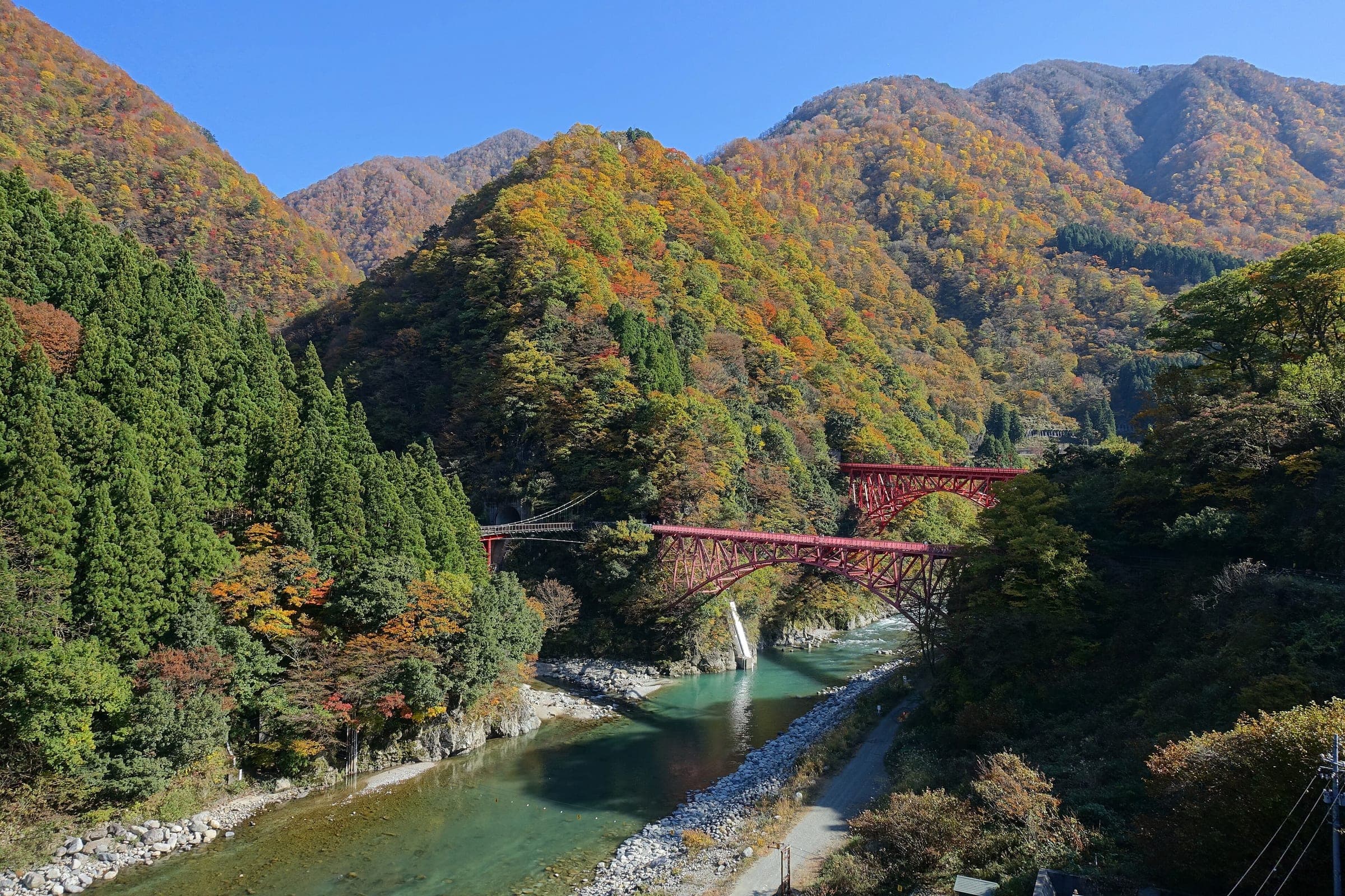 The red Yamabiko bridges span the jade Kurobe River below autumn mountains near Unazuki in Kurobe