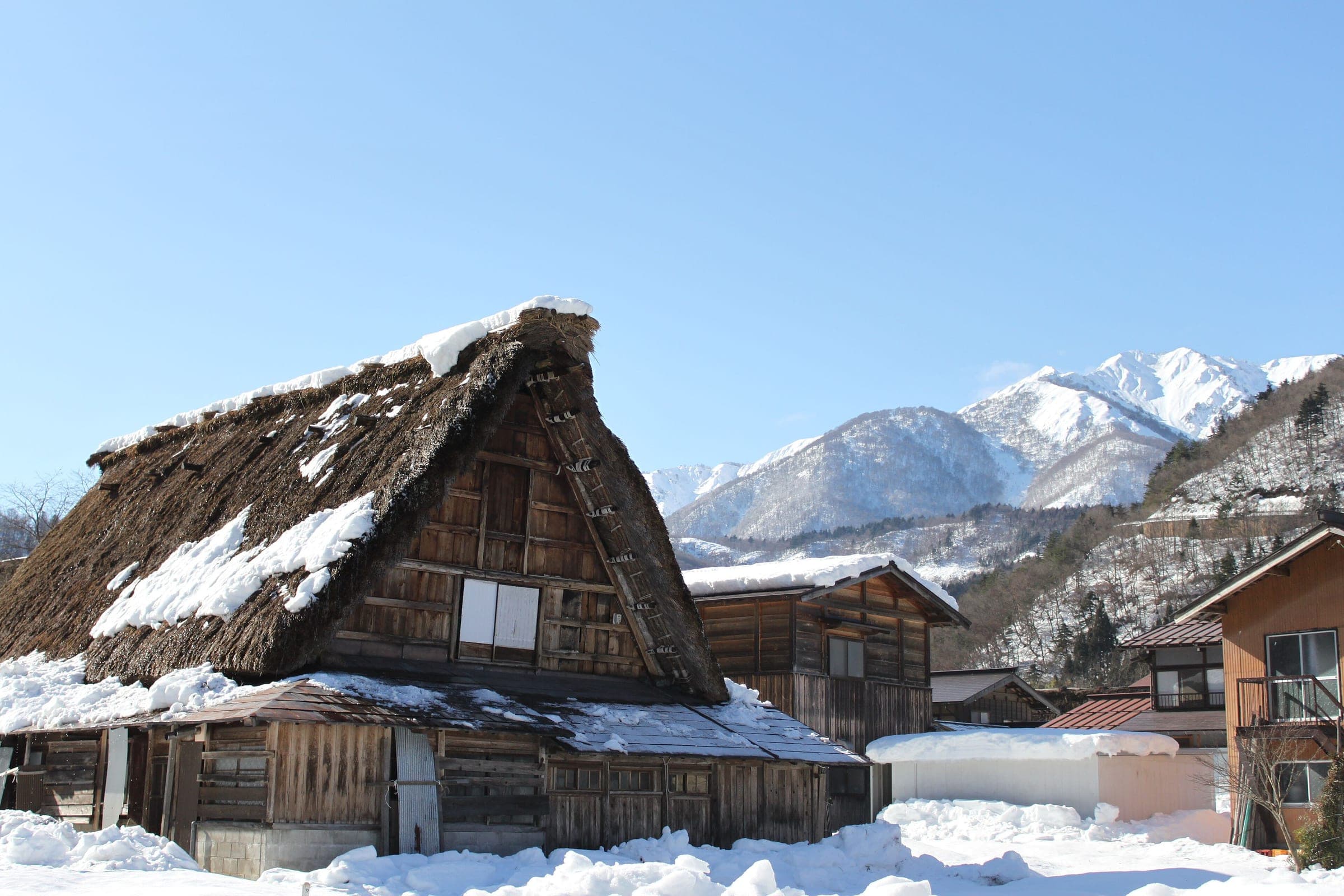 A thatched gassho-zukuri house in Shirakawa-go stands before snowy peaks under a clear blue sky